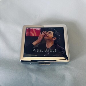 Puede incluir: Una cajetilla de cigarrillos plateada con una imagen colorida de una persona con el texto "Pills, Baby!" impreso en la parte delantera.