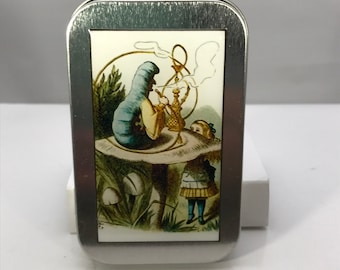 May include: A silver metal tin with a colorful image of Alice in Wonderland's caterpillar smoking a hookah on a mushroom with Alice standing nearby.