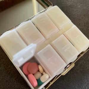 Puede incluir: Un organizador de pastillas de pl&aacute;stico blanco con siete compartimentos. Los compartimentos est&aacute;n etiquetados con los d&iacute;as de la semana. El organizador est&aacute; abierto y hay pastillas en uno de los compartimentos.