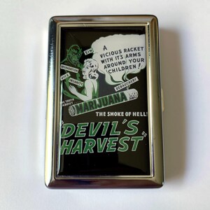 May include: A silver cigarette case with a black and green graphic design. The graphic features a woman and a devil with the text "MARIJUANA THE SMOKE OF HELL! DEVIL'S HARVEST".
