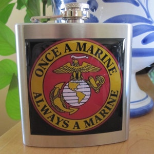 May include: A silver flask with a black and red sticker featuring the US Marine Corps emblem and the text "Once a Marine Always a Marine".