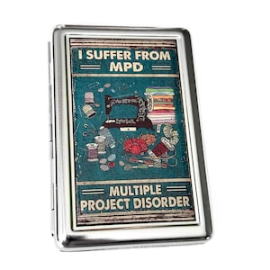 May include: A silver metal cigarette case with a colourful graphic design. The design features a sewing machine, thread spools, and fabric. The text on the case reads "I suffer from MPD - Multiple Project Disorder".