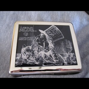 May include: A silver cigarette case with a black and white image of a character from Alice in Wonderland. The image has the text "MEDS... OR MADNESS" and "In this style 1906".