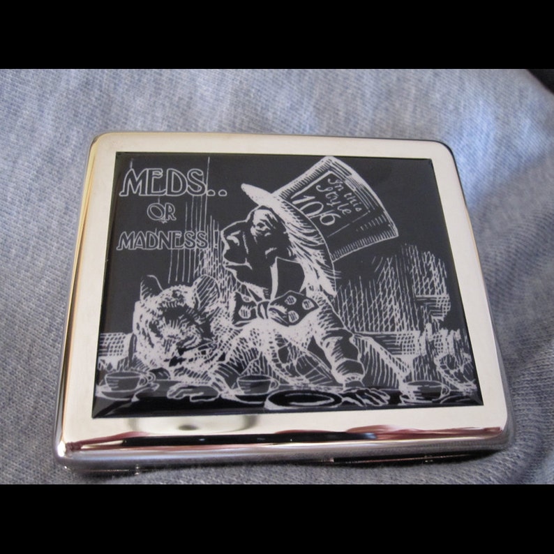 May include: A silver cigarette case with a black and white image of a character from Alice in Wonderland. The image has the text "MEDS... OR MADNESS" and "In this style 1906".