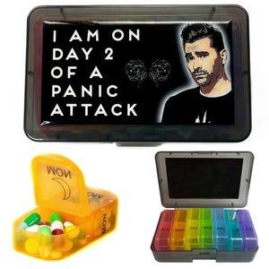 May include: A black pill box with a white graphic that says "I am on day 2 of a panic attack". The graphic also features a man with dark hair and a black shirt. The pill box is open and shows a yellow pill organizer with a moon graphic.
