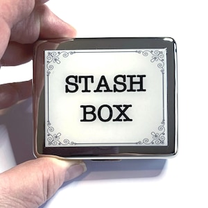 May include: A silver metal box with a white label that says "STASH BOX" in black lettering. The label has a decorative border.