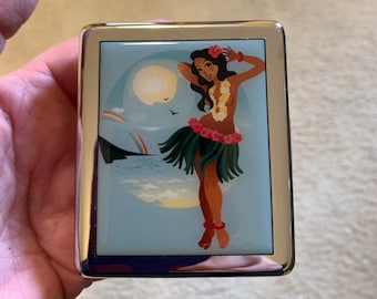 Pode incluir: Um estojo de cigarros prateado com uma imagem colorida de uma dançarina de hula em uma praia com um arco-íris e uma lua cheia.