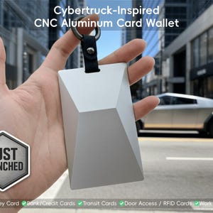 以下が含まれることがあります： サイバートラックのデザインにインスパイアされた、銀色のCNCアルミニウムカードウォレットが手に持たれています。ウォレットは幾何学的な形状で、黒いレザーストラップと金属リングが付いています。画像には「JUST LAUNCHED」と「Cybertruck-Inspired CNC Aluminum Card Wallet」と書かれています。