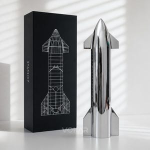 Könnte beinhalten: Ein glänzendes, silbernes Raketenmodell steht neben einer schwarzen Box. Die Box zeigt eine weiße Strichzeichnung der Rakete und das Wort "STARSHIP". Das Modell ist eine detaillierte Nachbildung eines Raumschiffs.