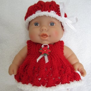 Puede incluir: Un vestido rojo de punto con ribetes blancos y un gorro rojo a juego de punto con ribetes blancos. El vestido tiene una cinta blanca y un broche de flor. La muñeca lleva polainas blancas de punto y zapatos rojos con lazos blancos.