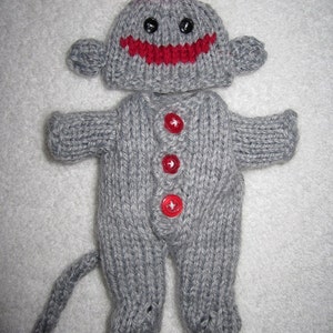 Puede incluir: Un mono de peluche gris tejido que lleva un gorro gris tejido con un pomp&oacute;n rojo y una sonrisa roja. La mu&ntilde;eca tiene cuatro botones rojos en la parte delantera de su mono tejido.
