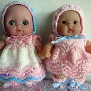 May include: Two baby dolls wearing pink and blue knitted dresses and hats. The dolls have large eyes and are smiling.