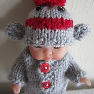 Puede incluir: Un gorro de mono de punto gris y rojo con un pomp&oacute;n rojo en la parte superior. El gorro tiene ojos de bot&oacute;n negros y un cuerpo de punto gris con botones rojos.