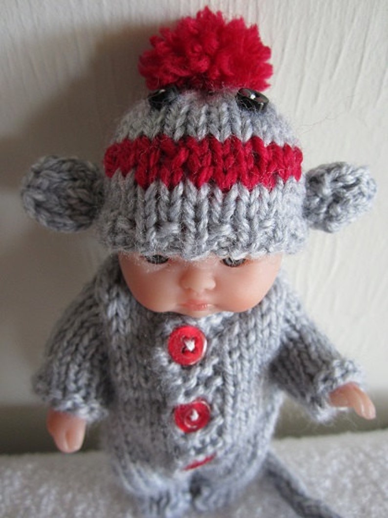 Puede incluir: Un gorro de mono de punto gris y rojo con un pomp&oacute;n rojo en la parte superior. El gorro tiene ojos de bot&oacute;n negros y un cuerpo de punto gris con botones rojos.