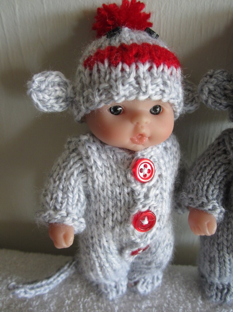 Puede incluir: Una mu&ntilde;eca de punto gris que lleva un gorro de punto gris y rojo con un pomp&oacute;n rojo. La mu&ntilde;eca tiene botones rojos en su atuendo.