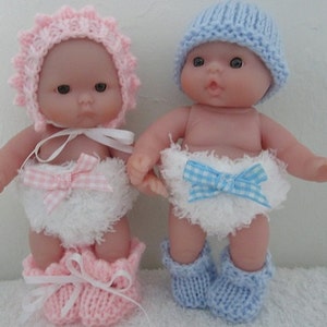 Könnte beinhalten: Zwei Baby-Puppen, die gestrickte Mützen und Stiefel tragen. Die Puppe links trägt eine rosa Mütze und Stiefel und eine weiße Windel mit einer rosa-weißen Karobinde. Die Puppe rechts trägt eine blaue Mütze und Stiefel und eine weiße Windel mit einer blau-weißen Karobinde.