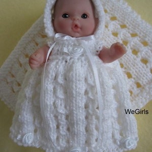 May include: A white crocheted baby doll dress with a hood and white satin ribbon. The dress has white fabric rosettes on the bodice and hem. The doll is wearing the dress and is lying on a white crocheted blanket.