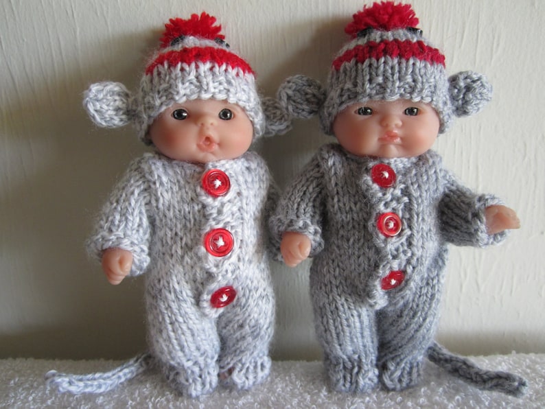 Puede incluir: Dos conjuntos de mu&ntilde;eca mono de punto gris con gorros a rayas rojas y blancas. Los conjuntos tienen botones rojos y una cola.