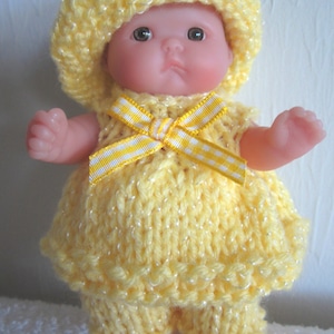 May include: A knitted outfit for a baby doll, in yellow with a white and yellow striped bow. The outfit includes a hat, dress, and trousers.