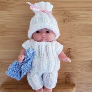 May include: A small doll wearing a white knitted hat with pink and blue accents and a white knitted outfit with pink and blue speckles. The doll is holding a blue knitted blanket.