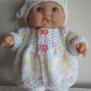 May include: A knitted baby doll outfit in a pastel color scheme with a white hat and pink buttons. The outfit includes a cardigan, pants, and booties.