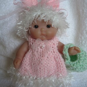 May include: A small doll wearing a pink knitted dress with white fur trim, a white fur hat, and pink bunny ears. The doll is holding a green knitted basket.