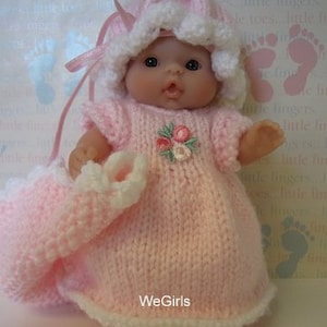 Puede incluir: Un vestido de muñeca de bebé de punto rosa con ribetes blancos y un diseño de bordado floral. La muñeca lleva un gorro de punto rosa a juego con ribetes blancos.
