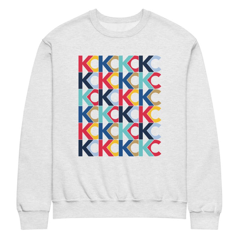 May include: A light grey sweatshirt with a repeating pattern of the letters "K" and "C" in various colours, including red, blue, yellow, and brown. The design is a geometric arrangement of the letters.