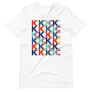 Puede incluir: Camiseta blanca con un patrón geométrico de las letras "K" y "C" en rojo, azul, amarillo y verde.
