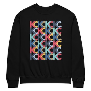 May include: Black sweatshirt featuring a repeating pattern of the letters "KC" in a variety of colours, including red, blue, yellow, and teal. The letters are arranged in a grid-like design across the front of the garment.