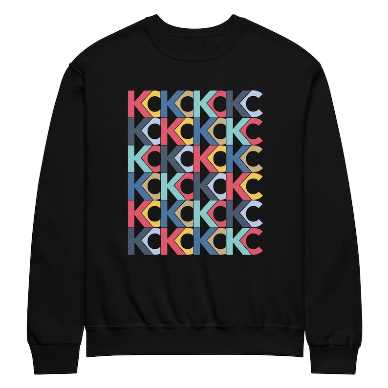 May include: Black sweatshirt featuring a repeating pattern of the letters "KC" in a variety of colours, including red, blue, yellow, and teal. The letters are arranged in a grid-like design across the front of the garment.