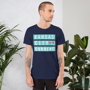 May include: A navy blue t-shirt with a light blue rectangle featuring the text "KANSAS CITY**" with two red stars and the text "CURRENT" in white.