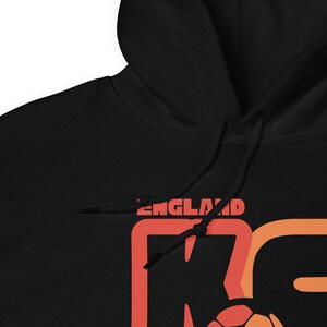 May include: Black hooded sweatshirt with the word "ENGLAND" in red letters. The sweatshirt has a large graphic in orange and red, with a soccer ball design. The hood has drawstrings.