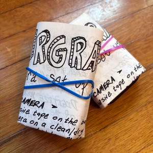 May include: Two white packages wrapped in paper with black marker writing. The packages are tied with rubber bands. The writing on the packages says "CAMERA - adhesive tape on the" and "RGRA ra sat".