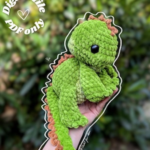 May include: A green crocheted dinosaur with brown accents. The plush toy has a textured, fuzzy appearance and large black eyes. The image includes the text "Digital file PDF only".