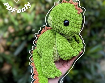 May include: A green crocheted dinosaur with brown accents. The plush toy has a textured, fuzzy appearance and large black eyes. The image includes the text "Digital file PDF only".