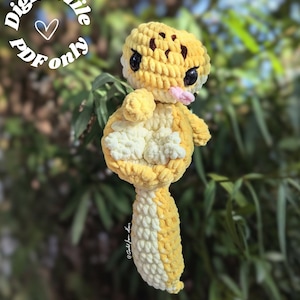 Puede incluir: Un peluche de gecko tejido a crochet en amarillo y blanco, con ojos negros y una lengua rosa. El gecko tiene manchas marrones en la cabeza y cuelga de una rama verde. El texto en un círculo dice "Digital file PDF only".