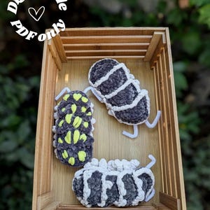Peut inclure: Trois cloportes en peluche crochetés dans un plateau en bois. Deux sont gris avec des détails blancs et un est gris avec des taches jaunes. Le texte "Digital file PDF only" est disposé en cercle en haut à gauche, avec un cœur.