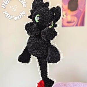 May include: A black crochet dragon plushie with green eyes and a red tail. The dragon is made of a soft, textured yarn. The text "Digital file PDF only" is in a white circle above the dragon.