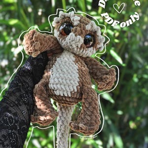 May include: A brown and cream-colored crocheted plush creature with large dark eyes and a long tail. The plush has a textured, fuzzy appearance and is outlined in white. Text in the upper right corner reads Digital file PDF only.