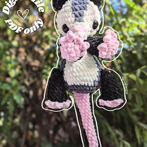 May include: A handmade crochet possum plushie in black, white, gray, and pink. The plushie has a long pink tail and is holding its pink paws up. The text "Digital file PDF only" is in a circle above the possum.
