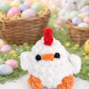 Puede incluir: Un pollo blanco de ganchillo con cresta roja, pico y patas naranjas y ojos negros. El pollo está sobre hierba verde, con huevos de Pascua y cestas en el fondo.