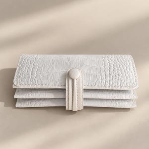 May include: A rectangular off-white wallet featuring a textured crocodile skin pattern. The wallet has a tri-fold design with multiple horizontal compartments and is secured in the centre by a vertical strap with a matching circular button closure.
