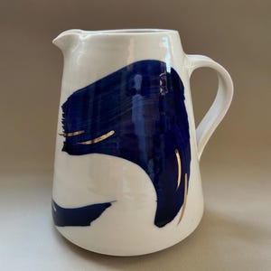 Puede incluir: Jarra de cerámica blanca con asa y boquilla curvadas. La jarra está decorada con un gran diseño abstracto de pincelada azul y detalles dorados. Una pieza decorativa para la cocina o el comedor.