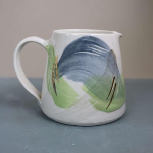 Puede incluir: Jarra de cerámica blanca con asa curva y boquilla. La jarra está decorada con pinceladas abstractas en tonos azules, verdes y dorados. El diseño se aplica en la parte delantera de la jarra.