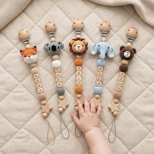 May include: Five pacifier clips featuring crocheted animal heads including a fox, koala, lion, elephant, and bear. Each clip has wooden beads, some with letters spelling NORA, THEO, ARLO, MILA, and FINN, and a wooden star charm at the bottom.