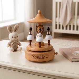 May include: A wooden musical carousel featuring three ballerina figures with dark brown bodies and white skirts. The base is engraved with the name Sophie and a heart symbol. A small wooden knob on the side serves as the winding mechanism for the music.