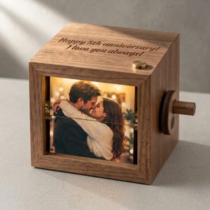 May include: A square wooden music box featuring a photo of a couple embracing on the front panel. The top surface is engraved with the text Happy 5th anniversary! I love you always! A small circular wooden crank handle is attached to the right side.