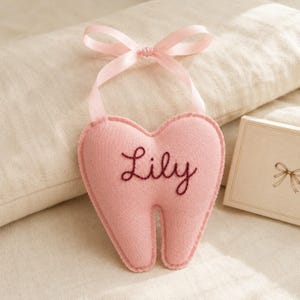 May include: A soft, light pink felt tooth-shaped pillow featuring the name Lily embroidered in dark maroon thread across the center. The pillow is topped with a matching light pink satin ribbon tied into a bow for hanging purposes.