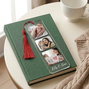 May include: Clear acrylic bookmark with three photo inserts and a red tassel. The bookmark rests on a green book and features the text "Lily & Ben". The photos show a baby's feet, a mother and child, and a drawing.
